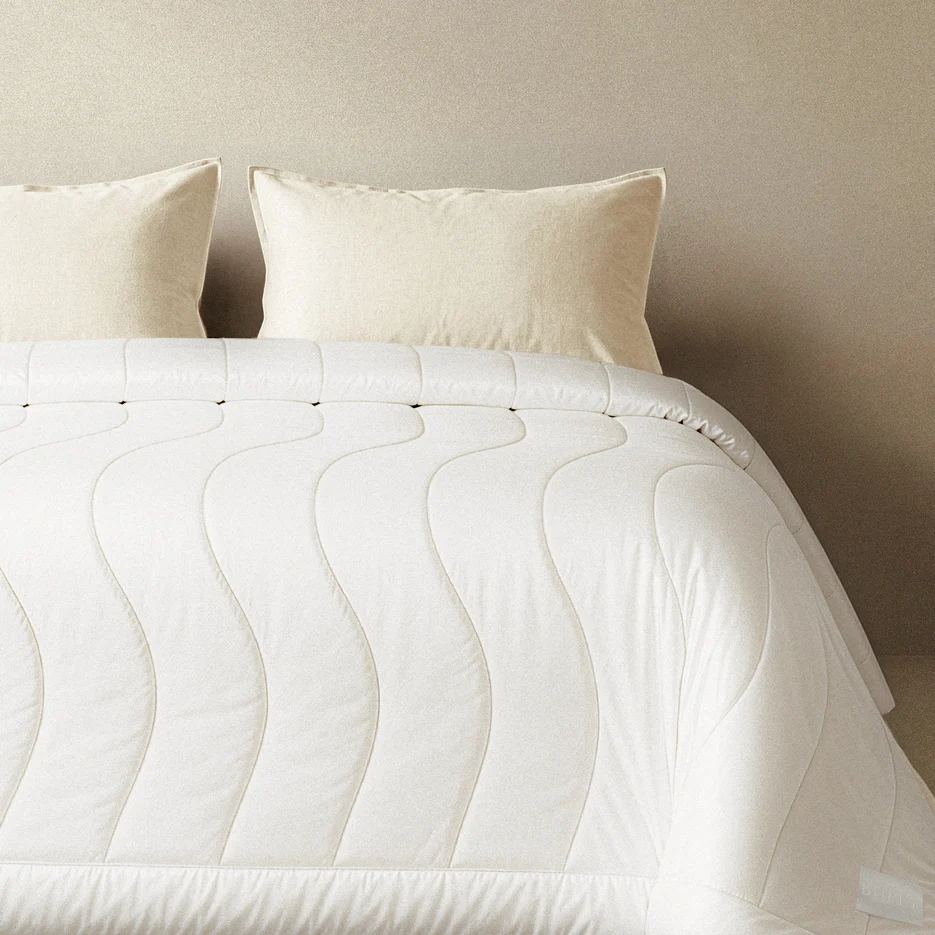 White Breeze Comforter laid over a bed, with two cream pillows against a warm beige wall.